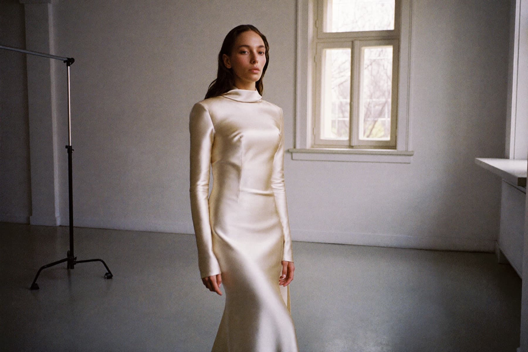 Une mariée en robe de soie ivoire, seule dans un atelier vide, devant une fenêtre haute